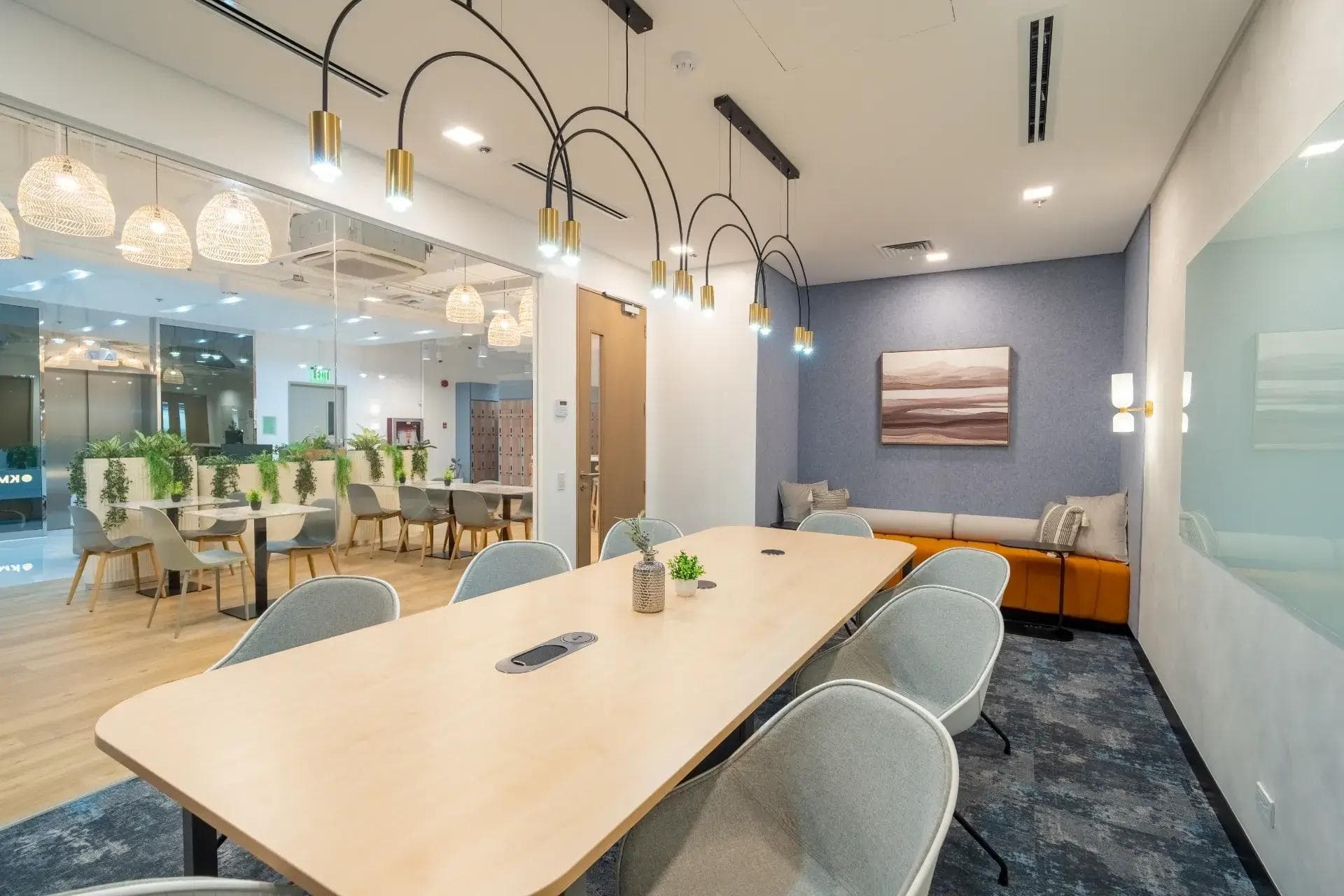 Glass-walled meeting room with long table, arch lights, sofa nook, and view of lounge with pendant lamps and indoor greenery.