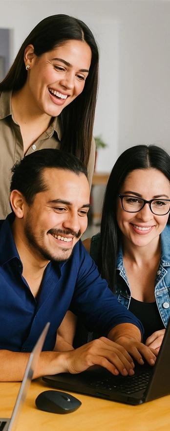 Three colleagues smiling and collaborating closely over a laptop in a casual workspace setting.