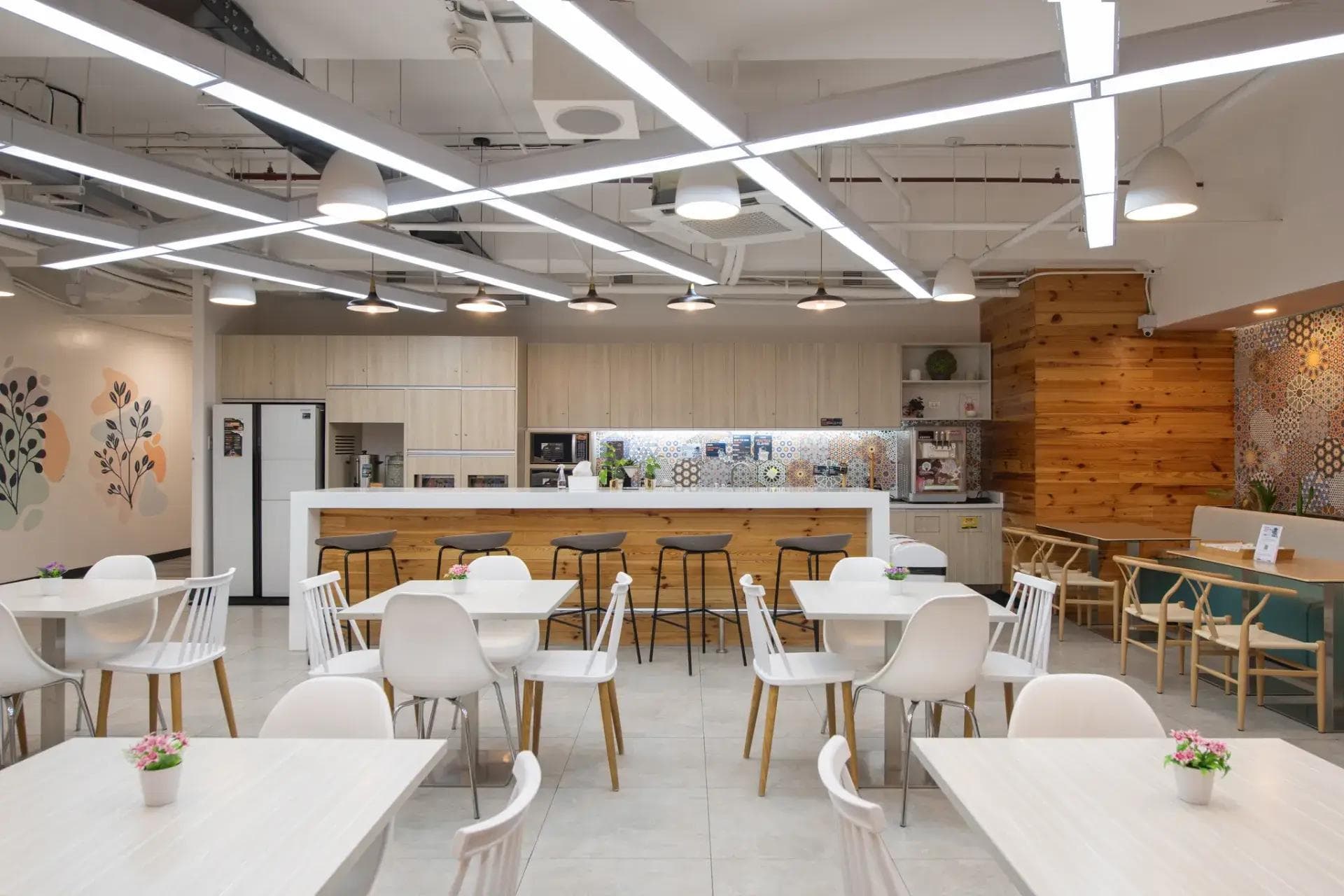 Modern pantry at KMC Mandaluyong office space with light wood cabinets, bar stools, white tables, and floral wall accents.