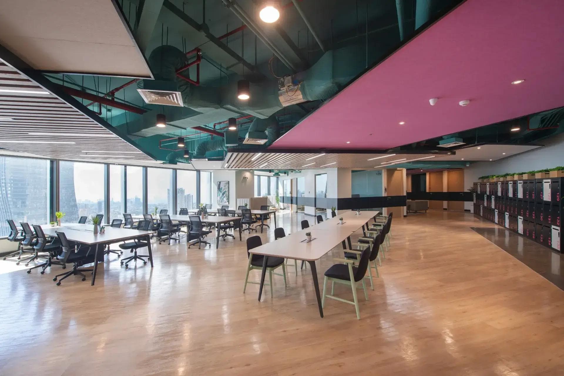 Open-plan coworking space with hot desks, ergonomic chairs, lockers, and panoramic windows offering city skyline views.