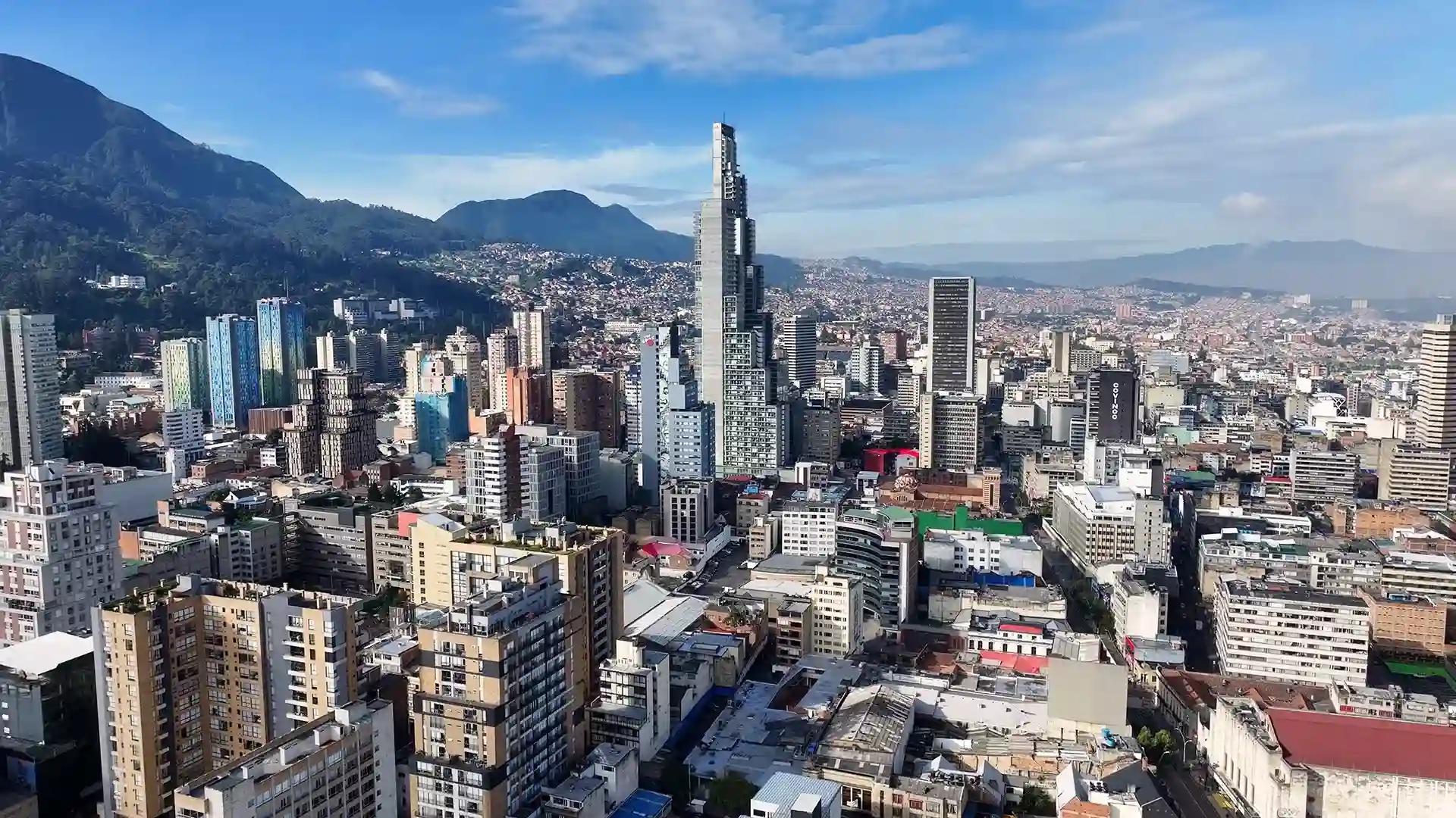 Aerial view of Bogotá with mountain backdrop, dense urban buildings, and modern high-rise towers under a clear blue sky.