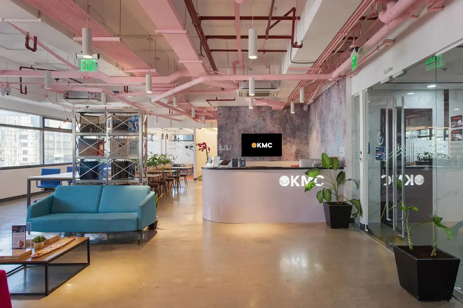 A modern KMC reception area with industrial-chic decor, pink ceilings, cozy seating, and lush greenery decorations.