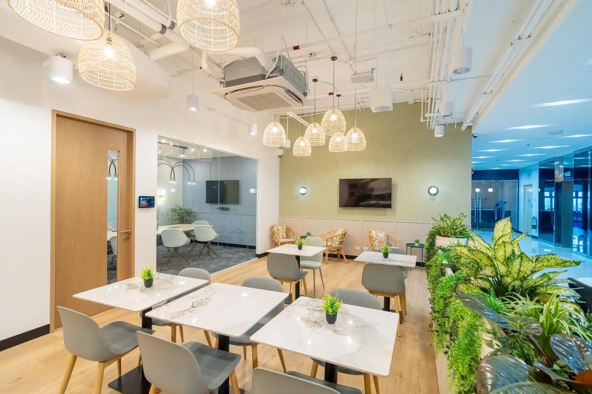 Bright lounge with marble tables, rattan chairs, pendant lights, glass room and lush indoor greenery along a planter wall.