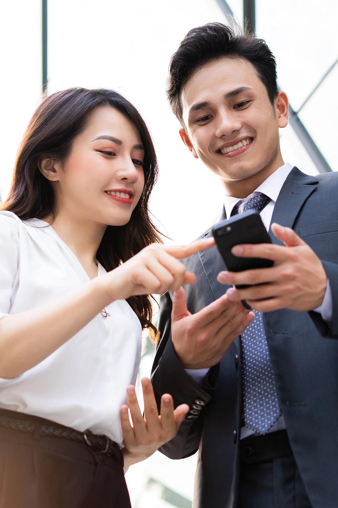 Two professionals smiling while looking at a smartphone outside a modern glass office building.