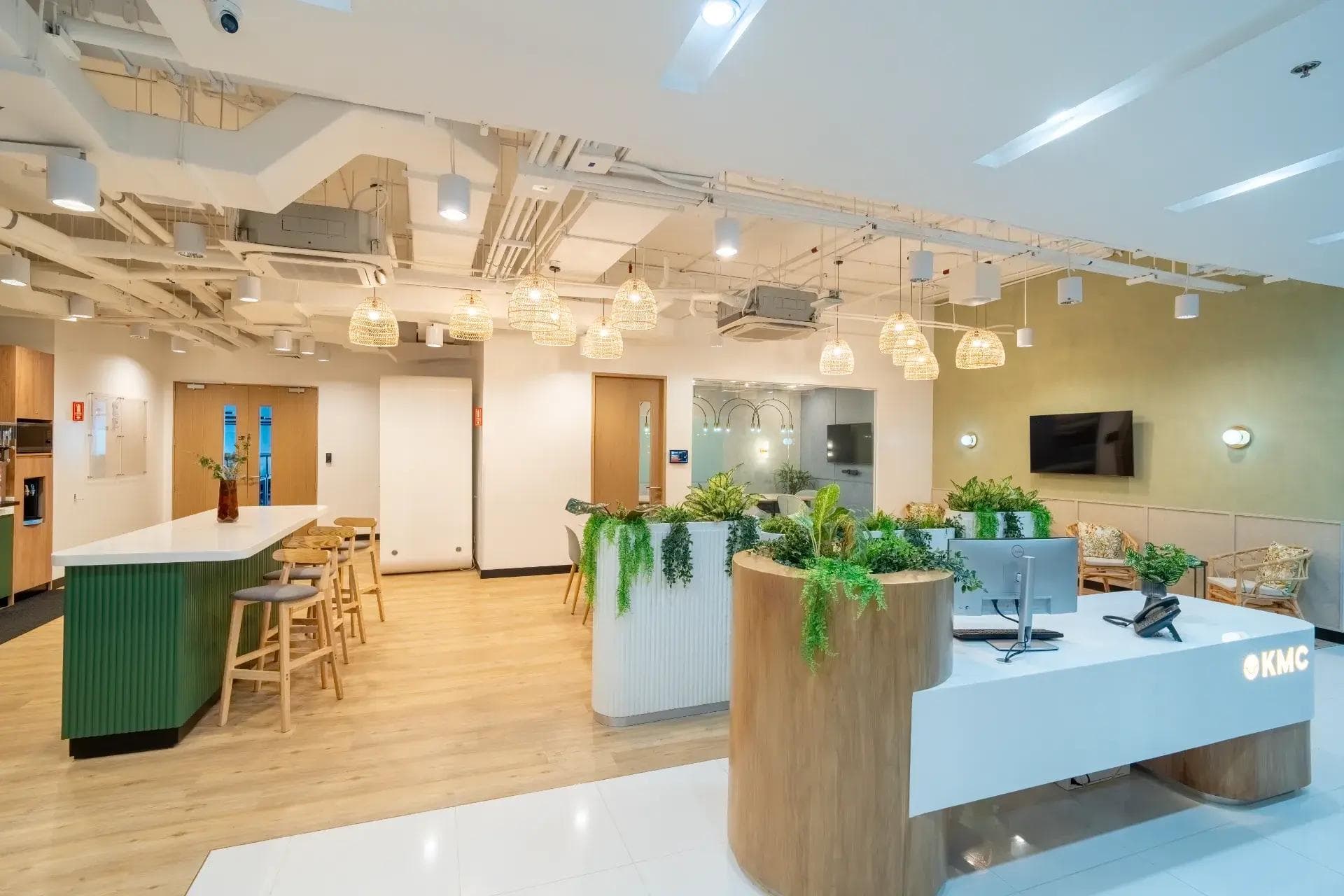 Bright coworking reception with white counter, wood floors, pendant lights, open ceiling, and lush indoor plants & greenery.