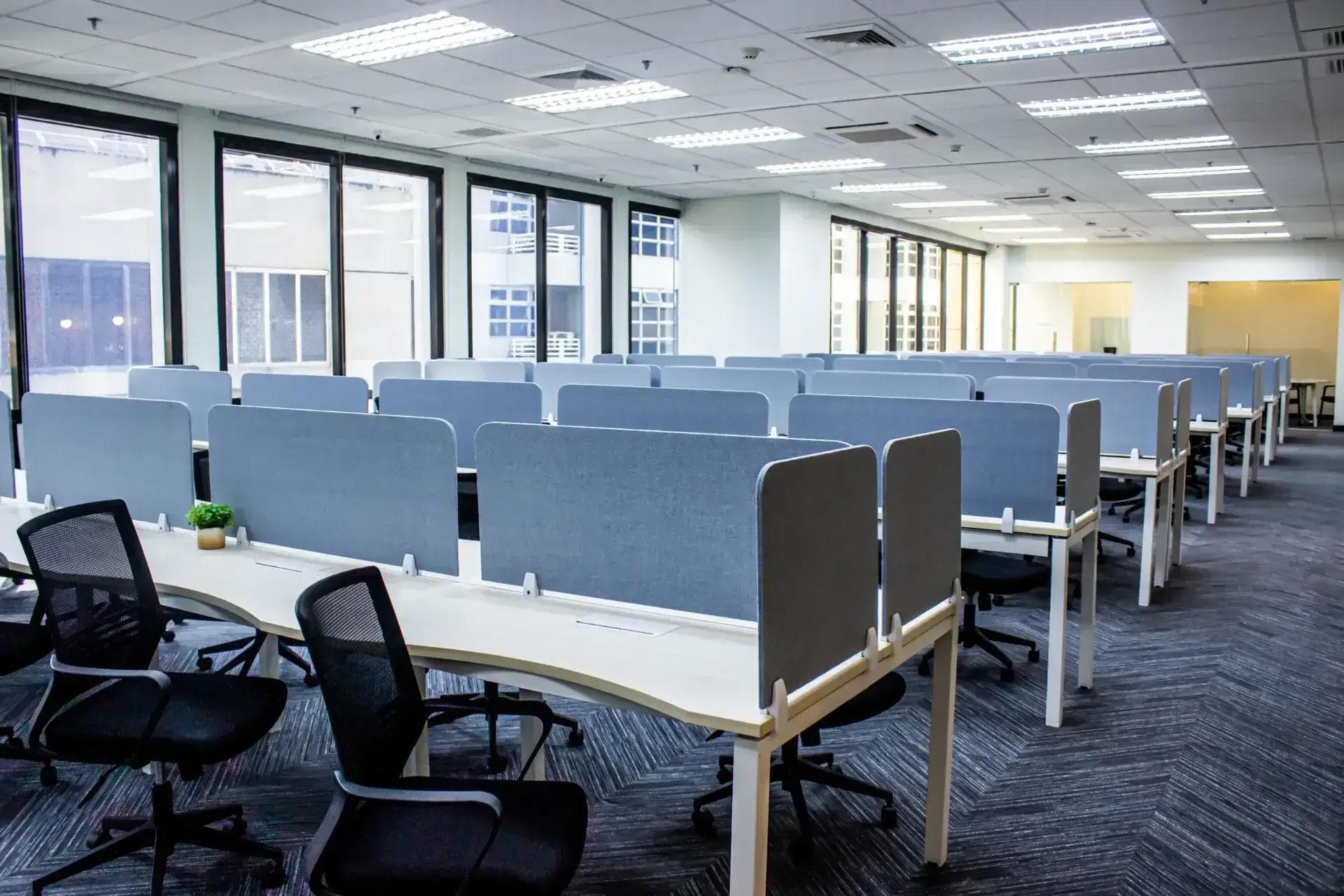 Spacious office floor with ergonomic chairs and partitioned desks, surrounded by tall windows letting in ample natural light.
