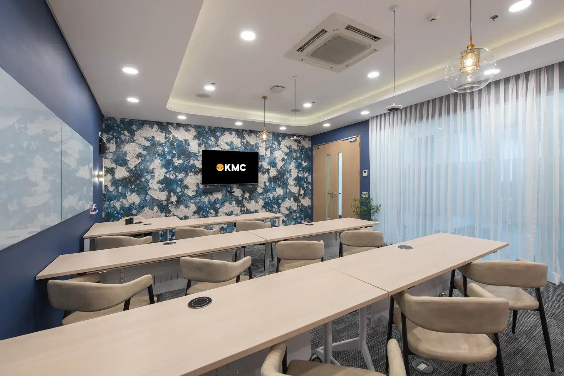 A training room with sleek white desks, beige chairs, a KMC logo shown on a TV screen, and a stylish blue-themed interior.